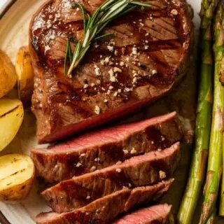 Top-down image of Bourbon Glazed Grilled Steak, plated at the center of a round ceramic dish. The steak is coated in a rich, caramelized bourbon glaze with perfect grill marks. Garnished with fresh rosemary and cracked black pepper, this smoky, juicy steak is served with roasted potatoes and asparagus for the ultimate steak dinner.