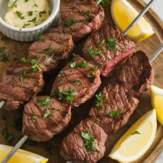 Top-down image of Garlic Butter Steak Skewers served on a wooden board. The tender, juicy steak pieces are coated in garlic butter and garnished with fresh parsley. A side of extra melted butter and lemon wedges complete this flavorful, easy-to-make dish.