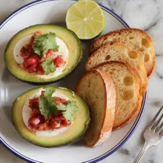 A photo of a serving plate with two baked avocado egg halves topped with chunky salsa and sprinkled with fresh cilantro. A lime wedge rests beside the avocados, and toast slices are arranged neatly next to the plate. Everything is placed on a marble counter with a fork off to the side.