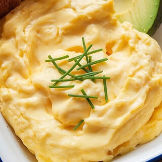 A photo of a white ceramic dish containing creamy scrambled eggs. The eggs are soft, glossy, and slightly rippled, showcasing their delicate texture. They are topped with a generous sprinkle of finely chopped chives. A slice of toast and sliced avocado are placed beside the eggs. The background is a wooden surface.