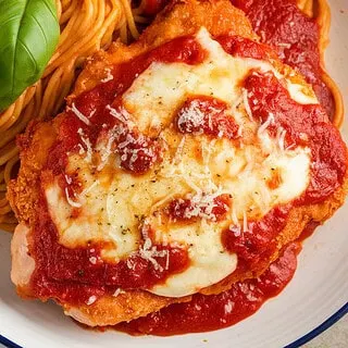 A photo of a plated serving of crispy chicken parmesan on a white ceramic plate. The chicken is topped with marinara sauce, melted mozzarella, and freshly grated parmesan. The dish is garnished with fresh basil leaves and served alongside spaghetti coated in marinara sauce.