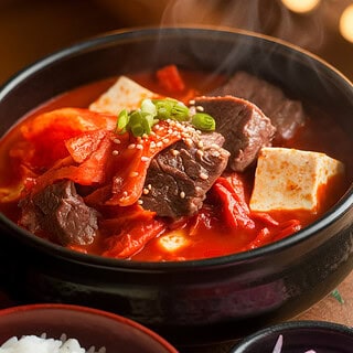 A bowl of Korean kimchi beef stew with beef, tofu, and mushrooms in a spicy red broth, served with white rice and pickled radish on the side.