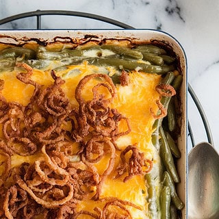 A photo of a golden, bubbling green bean casserole in a 9x13 dish, topped with crispy fried onions, fresh out of the oven. The dish sits on a trivet over a marble surface. A spoon rests beside it, ready to serve. The edges of the dish are slightly charred, showing the heat of the oven. The green beans are visible through the cheese layer, and the fried onions have a brown, crispy texture.