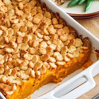 A photo of a freshly baked squash casserole in a white 9x13 dish with a golden cracker topping. The edges are bubbling slightly, and a serving spoon rests beside it. A portion is scooped out onto a plate next to roasted chicken and green beans.