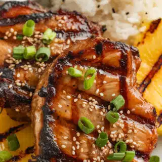 Grilled pineapple teriyaki pork chops with a caramelized glaze, served with coconut rice, grilled pineapple rings, and garnished with sesame seeds and green onions, with extra teriyaki sauce on the side.