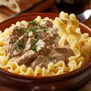 A bowl of Russian beef stroganoff served over buttery egg noodles, garnished with fresh parsley, with a side of rustic bread and a glass of red wine in the background.