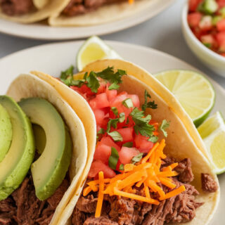 A photo of a plated serving of slow cooker beef tacos in warm corn tortillas. The tacos are filled with juicy, well-seasoned beef, topped with shredded cheese, chopped cilantro, diced tomatoes, and sliced avocado. Served with a wedge of lime and a side of pico de gallo.