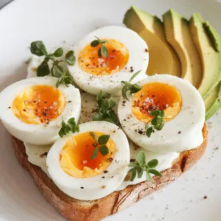A photo of a slice of toast topped with halved soft boiled eggs. The eggs are seasoned with salt and pepper and are garnished with fresh herbs. There is also a side of sliced avocado. The dish is plated on a white dish over a marble counter.