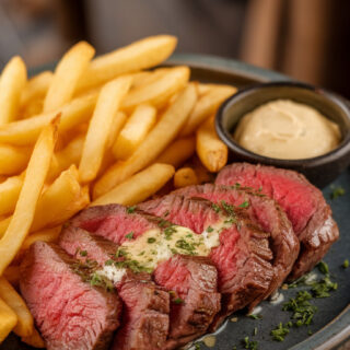 A photo of a finished plate of steak frites on a dark ceramic plate. The sliced steak is cooked medium-rare, with a pinkish-red center. It is drizzled with garlic butter and sprinkled with chopped parsley. The crispy golden fries are arranged neatly around the steak. A side of Dijon mustard and a small bowl of extra butter sit nearby for dipping. The background is blurred, showing a rustic setting with wooden elements.