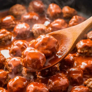 A skillet filled with Korean meatballs tossed in a thick, glossy gochujang sauce, with a wooden spoon stirring them for even coating.