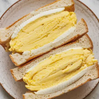 A top-view photo of the final serving of Tamago Sando on a white ceramic plate. The scrambled eggs are placed between soft slices of toasted white bread, with a light layer of mayonnaise. The sandwich is sliced and served on a marble countertop.