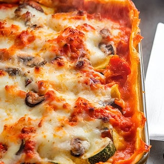 A photo of a golden, bubbling vegetable lasagna fresh out of the oven. The lasagna is filled with a medley of vegetables, including carrots, zucchini, and mushrooms. The top layer is a golden-brown, crispy layer of melted mozzarella and Parmesan cheese. The edges of the lasagna are slightly browned, and steam rises as the lasagna cools before slicing. A serving spatula is placed nearby, ready to plate.