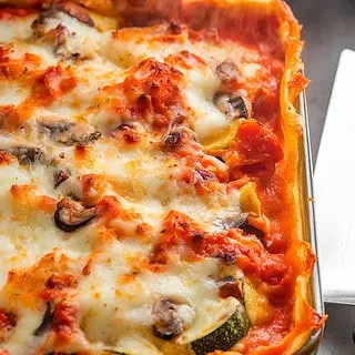 A photo of a golden, bubbling vegetable lasagna fresh out of the oven. The lasagna is filled with a medley of vegetables, including carrots, zucchini, and mushrooms. The top layer is a golden-brown, crispy layer of melted mozzarella and Parmesan cheese. The edges of the lasagna are slightly browned, and steam rises as the lasagna cools before slicing. A serving spatula is placed nearby, ready to plate.