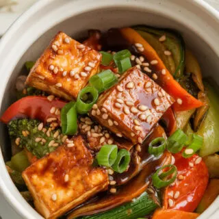 A photo of a white ceramic bowl filled with a vegetable stir-fry dish. The dish contains crispy tofu pieces, vibrant stir-fried vegetables, and a glossy, flavorful sauce. The bowl is garnished with sesame seeds and chopped green onions. There is a side of steamed rice. The background is clean.