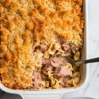 A photo of a freshly baked tuna noodle casserole in a white ceramic dish. The casserole has a golden breadcrumb topping and is filled with creamy noodles and chunks of tuna. A spoon is scooping out a portion of the casserole. The marble counter next to the casserole holds a plate with a serving of the casserole and a side of green salad.