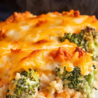 A photo of a final serving of cheesy broccoli-and-rice casserole. The cheese is melted and bubbling on top, and steam rises slightly from the cut edge. The dish is served on a white plate. There are green specks of broccoli and rice mixed with a creamy, orange sauce, topped with a golden, cheesy layer.