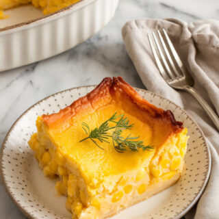 A photo of the final serving of corn pudding casserole. The edges are lightly browned and the center is soft and custardy. A fork and linen napkin sit beside the plate on a marble countertop.